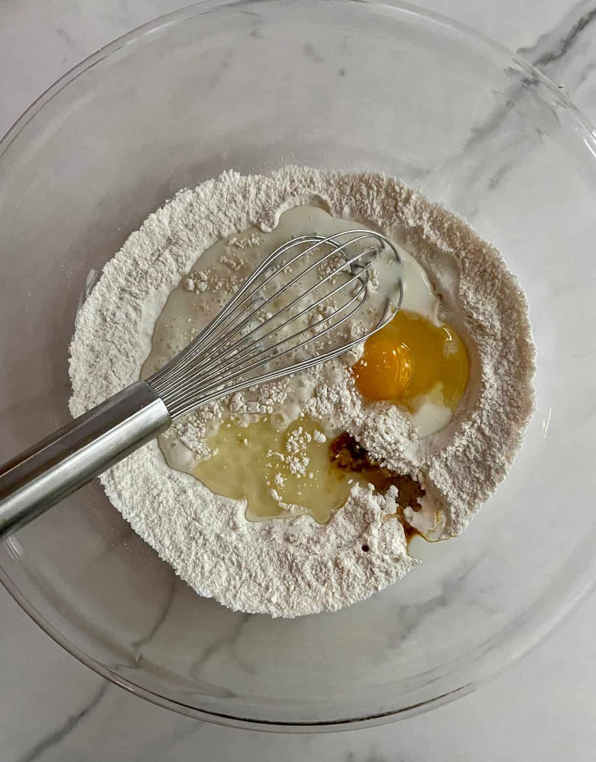 A large bowl containing flour, oil, an egg, baking powder, salt, vanilla, milk and sugar with a whisk.