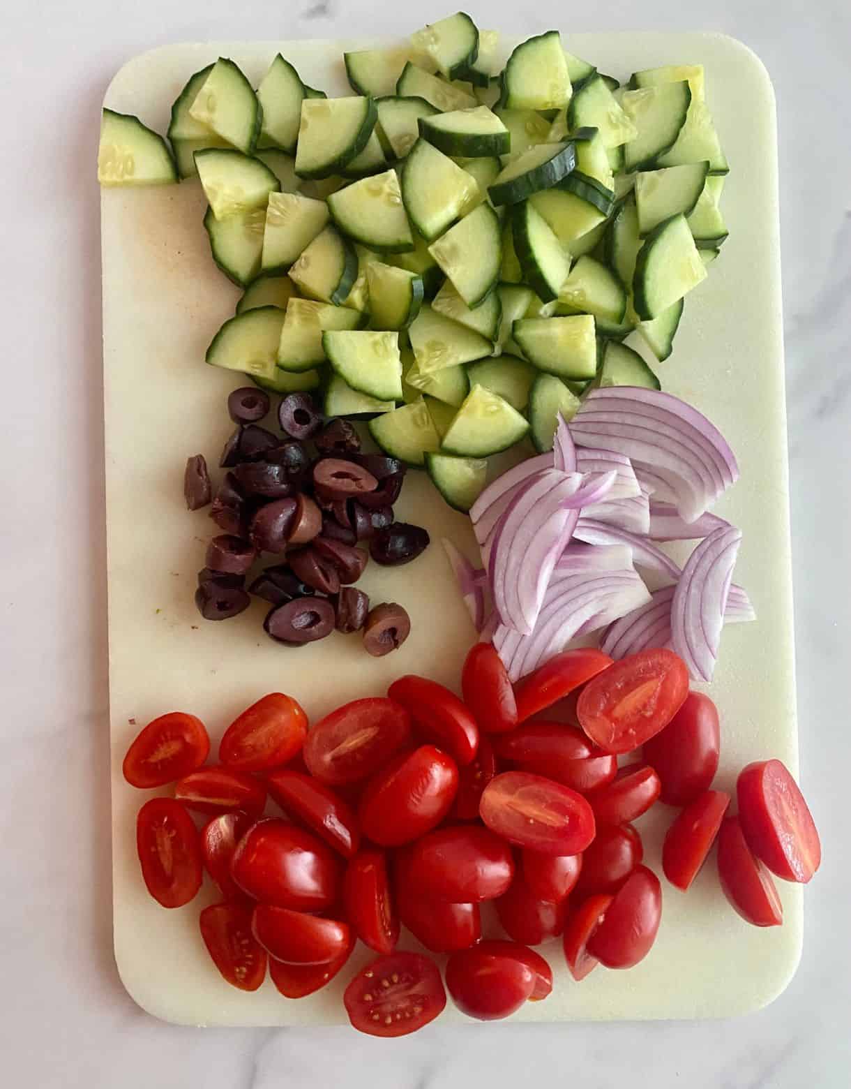 A cutting board with chopped cucumbers, sliced kalamata olives, sliced red onion and halved grape tomatoes for Chopped Greek Salad.