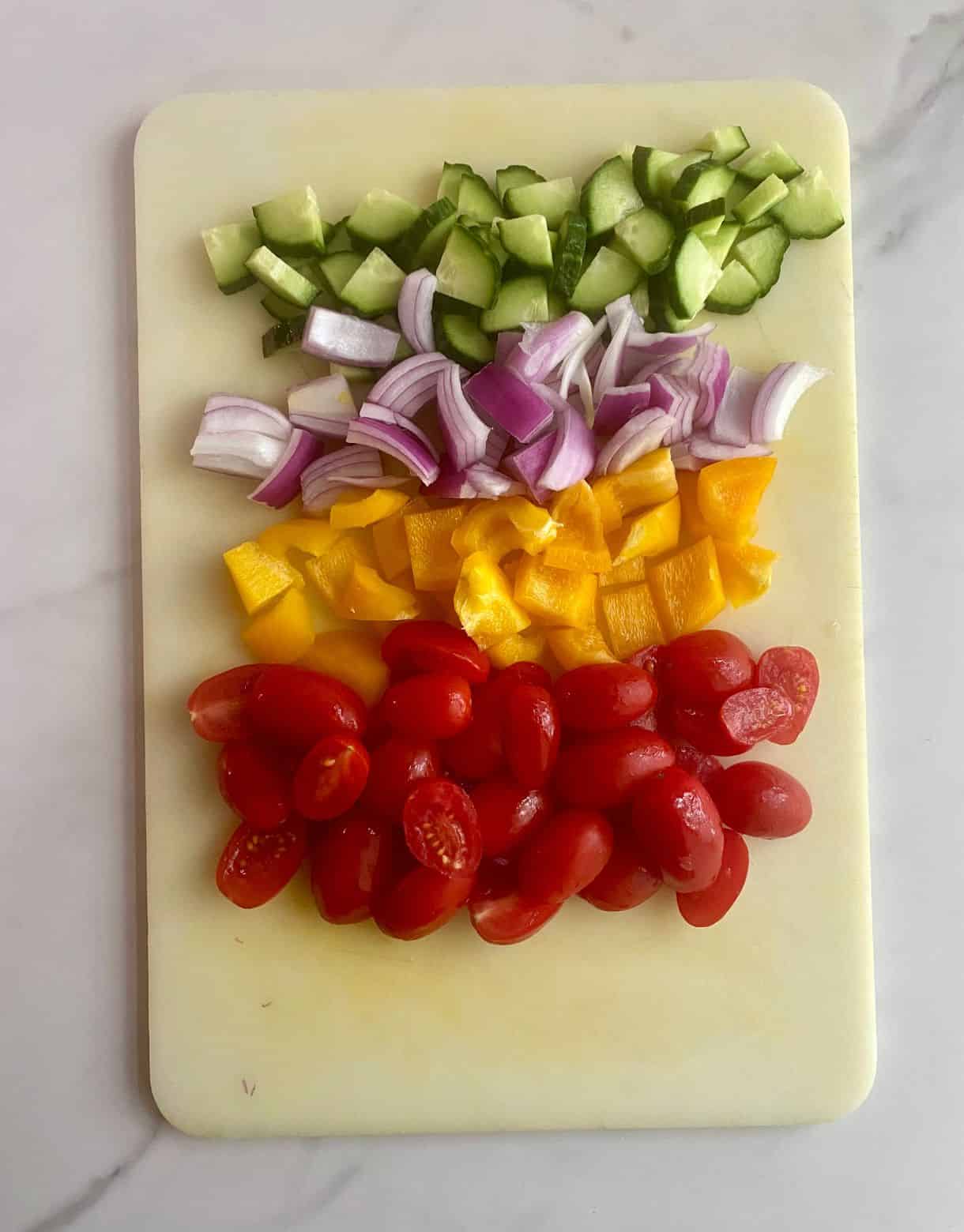 A cutting board with diced cucumbers, red onion, bell pepper and halved tomatoes.