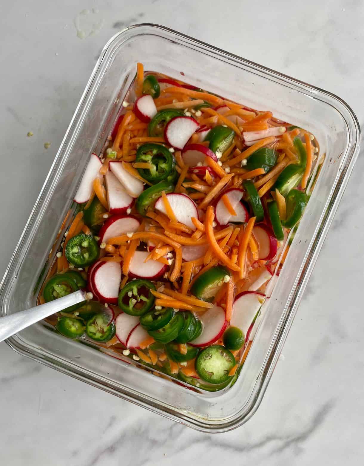A container of Vietnamese Pickled Vegetables with sliced jalapenos, sliced radishes and shredded carrots floating in vinegar.