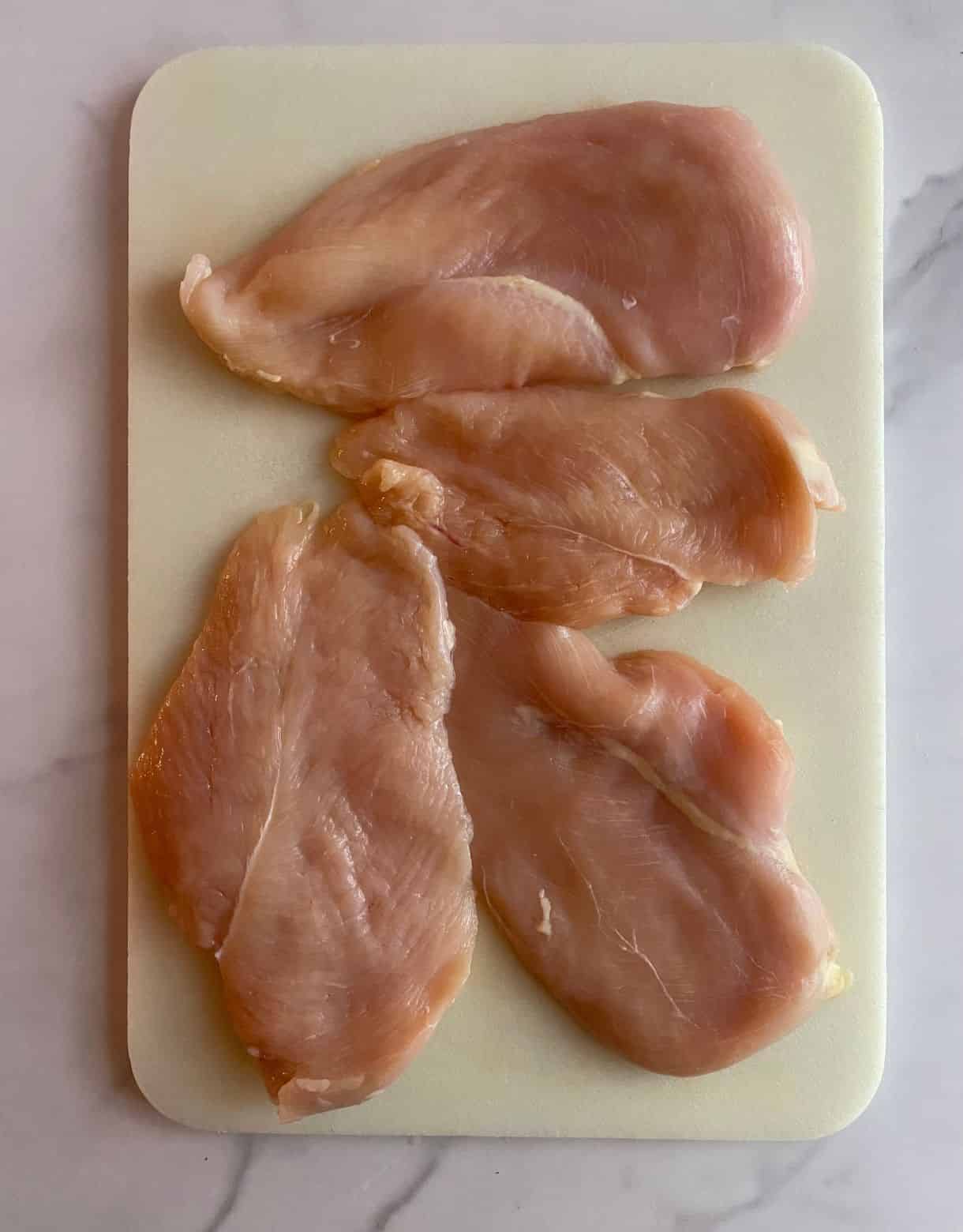 A cutting board with raw chicken breasts sliced in half lengthwise.