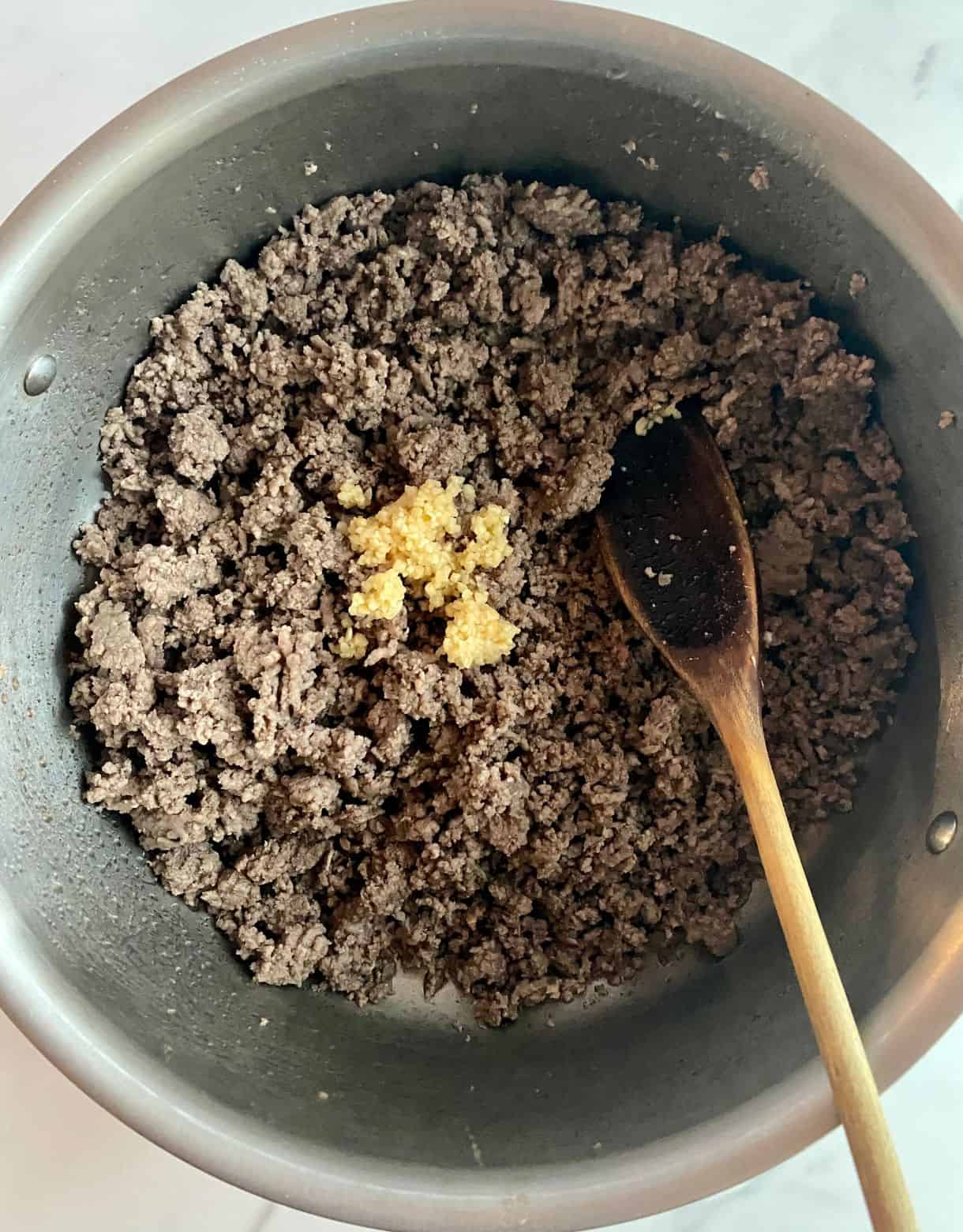 A skillet with cooked and crumbled ground beef and minced garlic added but not stirred.