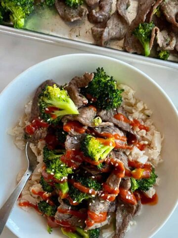 A bowl of Sheet Pan Beef and Broccoli over cauliflower rice and drizzled with sriracha sauce.
