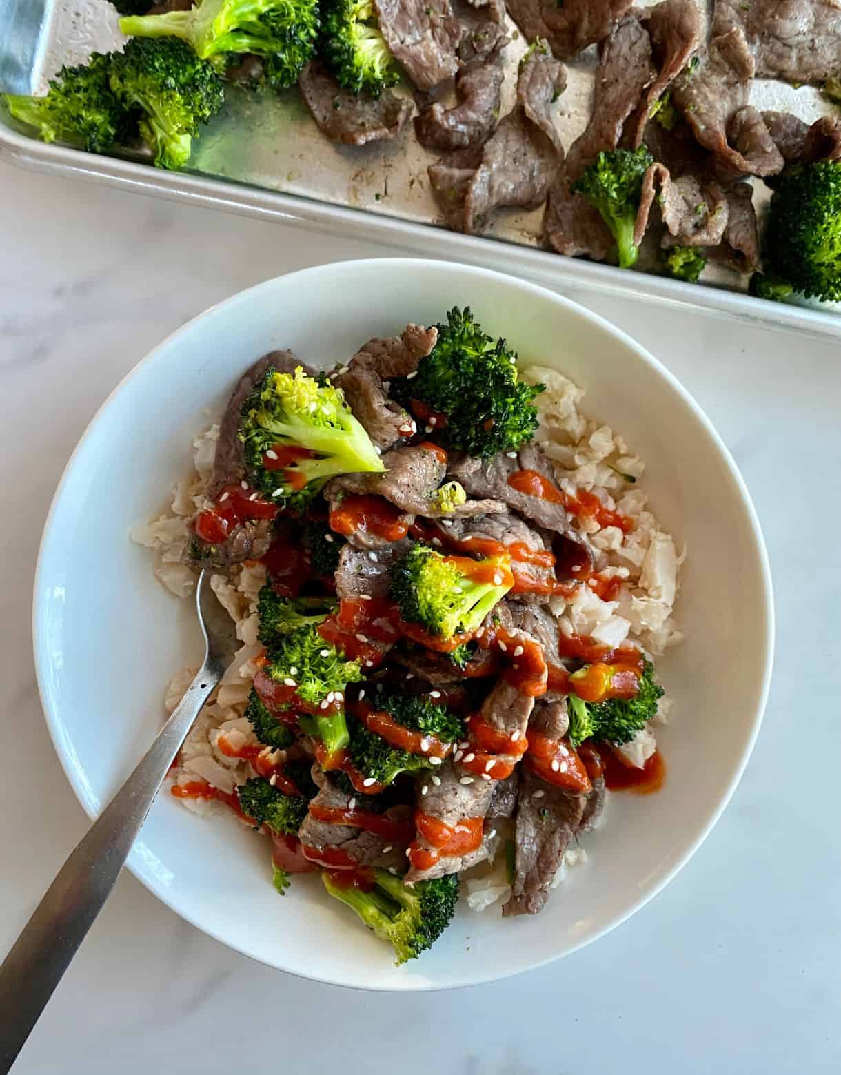 A bowl of Sheet Pan Beef and Broccoli over cauliflower rice and drizzled with sriracha sauce.