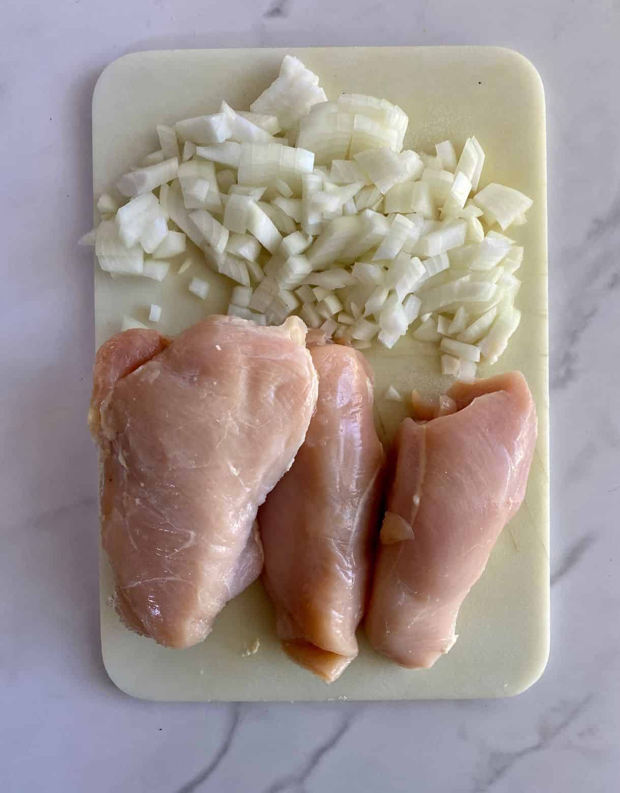 A cutting board with 3 trimmed chicken breasts and diced onion. 