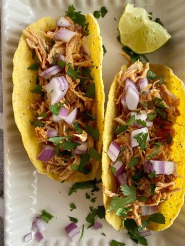 Two Slow Cooker Chicken Tacos on a plate with chopped raw and onion cilantro and a wedge of lime.