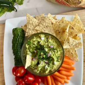 A plate with a bowl of Spicy Guacamole as well as sliced cucumbers, grape tomatoes, baby carrots and tortilla chips to dip in it.