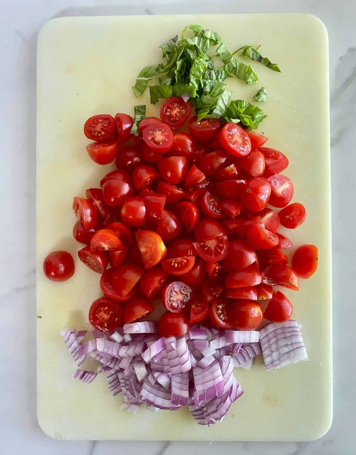 A cutting board with diced red onion, halved grape tomatoes and sliced basil.