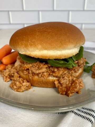 A bun with Buffalo Sloppy Joe topped with arugula on a plate with carrots.