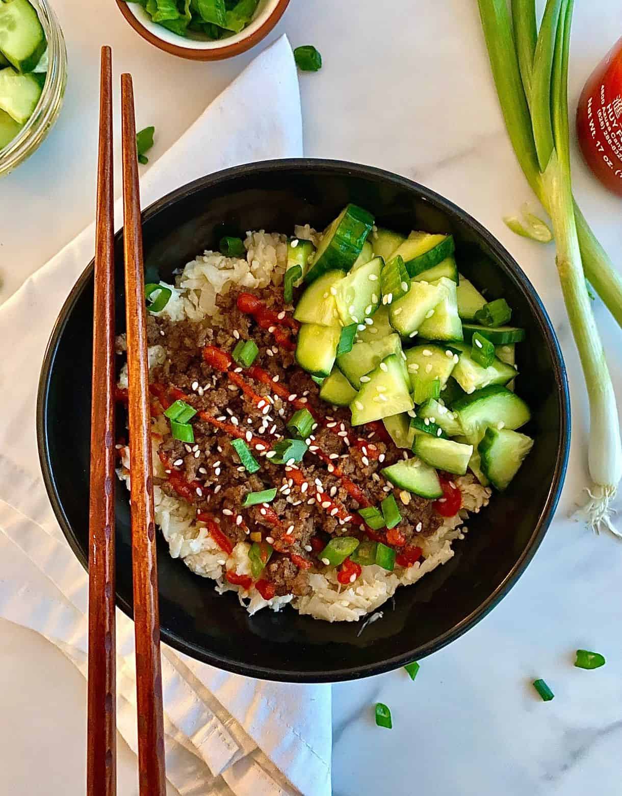 A Bulgogi Bowl with seasoned beef served over cauliflower rice and topped with sliced green onions, sesame seeds, pickled cucumbers and sriracha sauce.