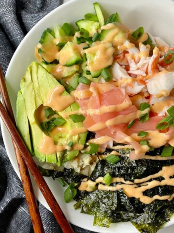A Deconstructed Sushi Bowl with rice, sliced avocado, sliced seaweed, crab, chopped cucumbers, sliced scallions and pickled ginger all topped with Sriracha Mayo.