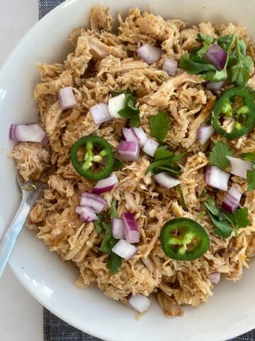 A bowl of cooked pork loin carnitas topped with sliced jalapenos and chopped onion and cilantro.