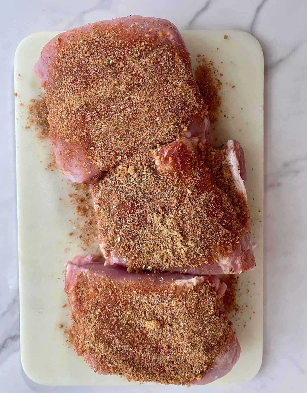 Three pieces of pork loin on a cutting board and rubbed with spices.