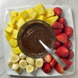 A plate with melted Chocolate Fondue surrounded by sliced pineapple, strawberries and bananas.