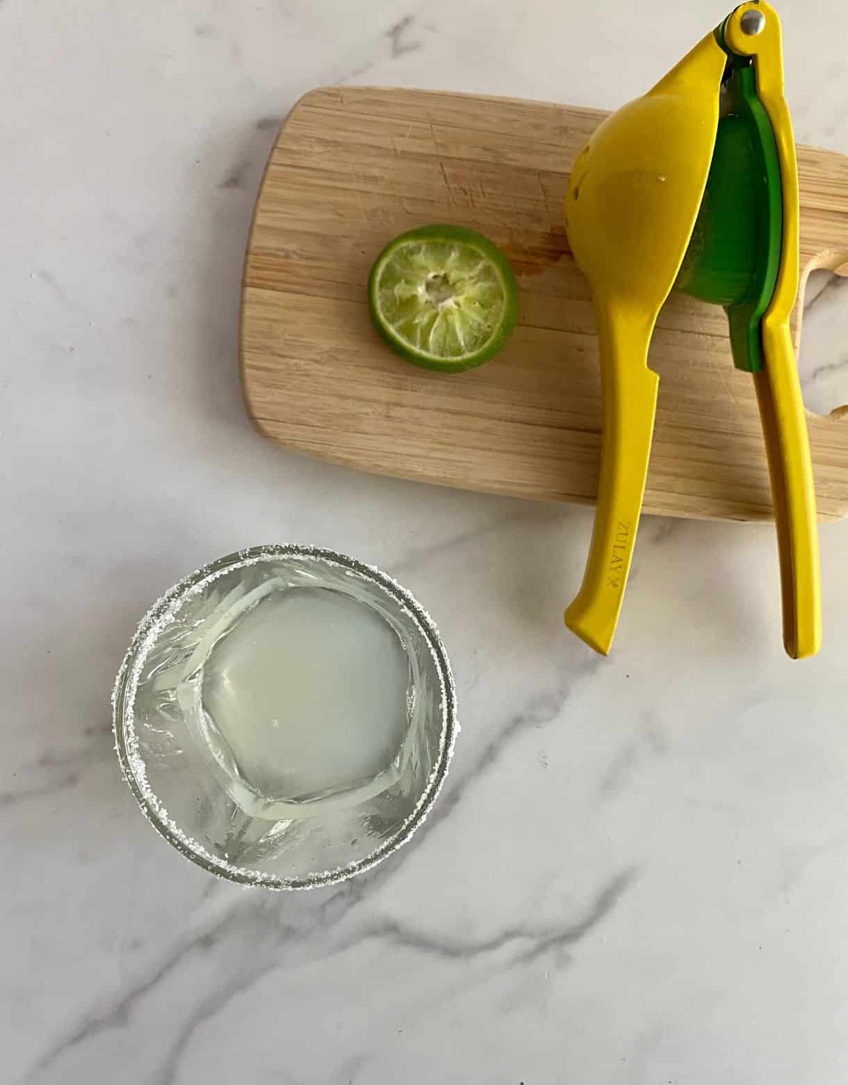 A glass with a salted rim with lime juice added. A cutting board with a juice press and a squeezed lime.