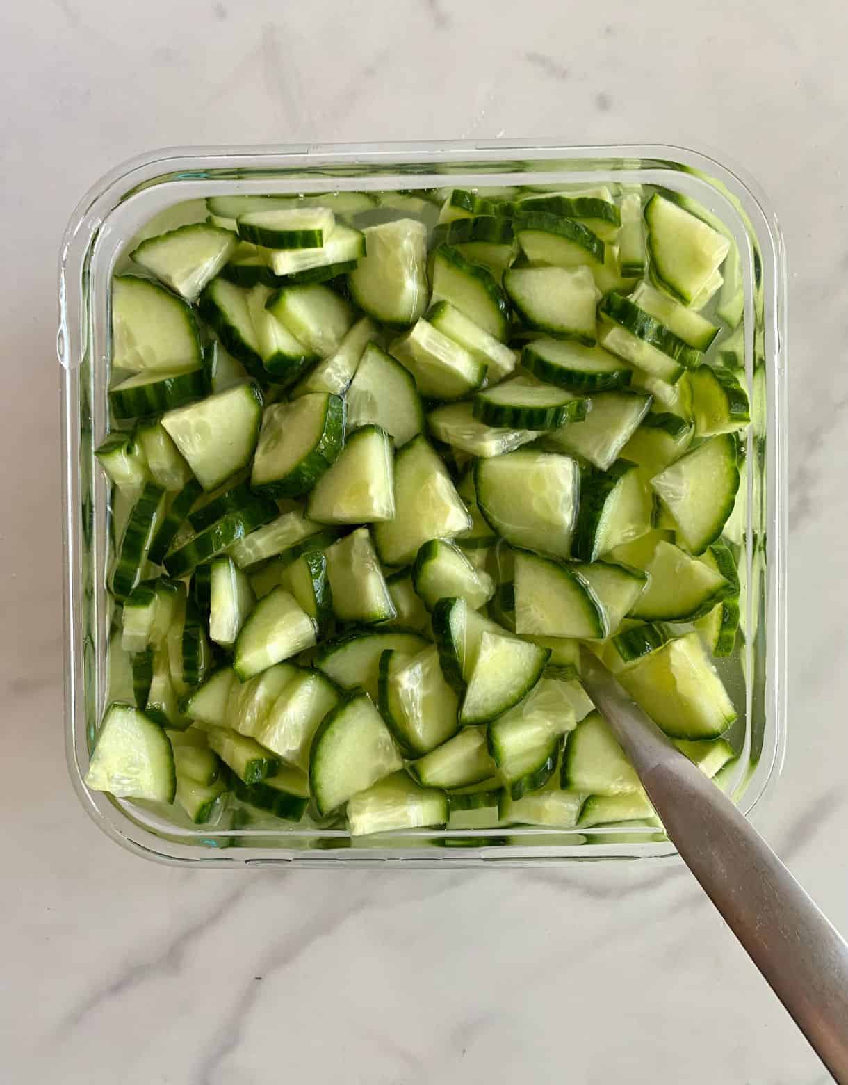 A glass dish with Pickled Cucumbers marinating in vinegar with a spoon.