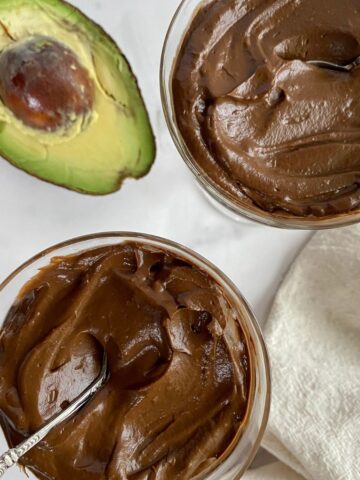 Two cups of Vegan Avocado Chocolate Mousse with spoons and half an avocado.