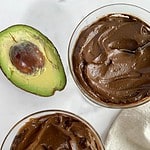 Two cups of Vegan Avocado Chocolate Mousse with spoons and half an avocado.