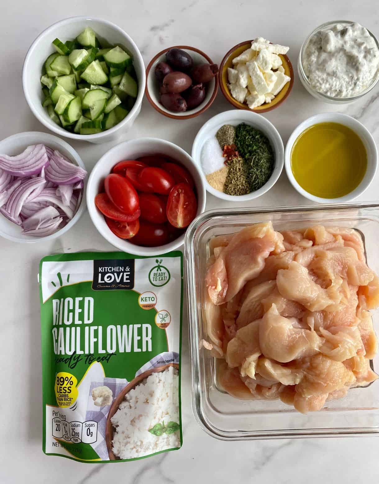 Sliced chicken breasts, a package of cauliflower rice, chopped cucumbers, red onion and tomatoes, olives, spices, feta cheese, olive and tzatziki sauce.