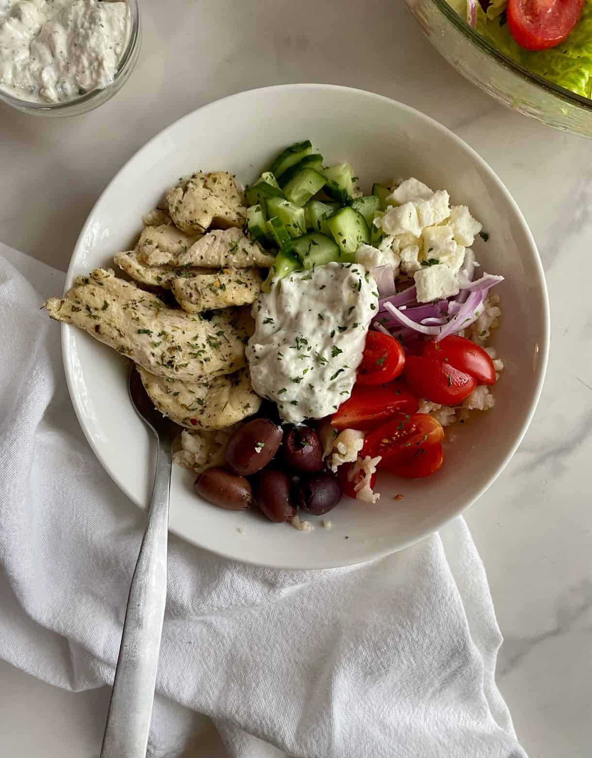 A Chicken Tzatziki Bowl with cauliflower rice, cooked chicken, olives, cucumbers, feta, tomatoes, red onion and tzatziki sauce.