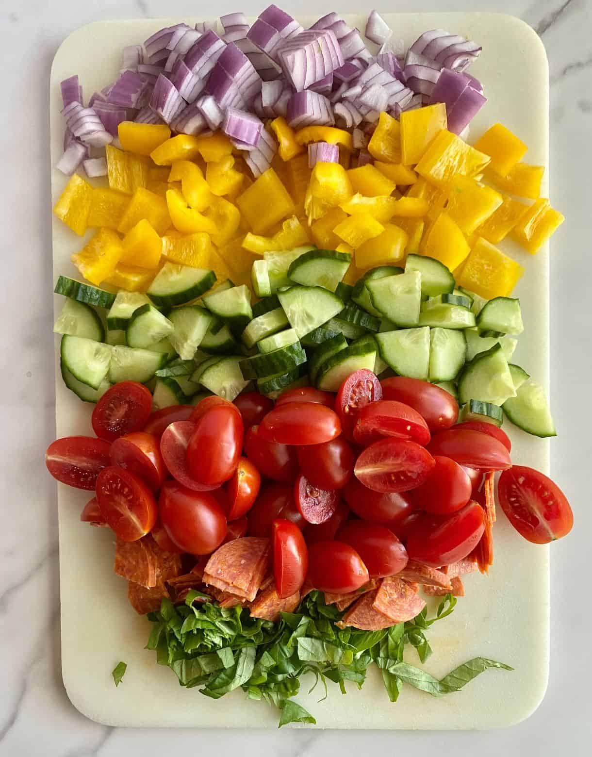 A cutting board with diced cucumbers, red onion, bell pepper, halved tomatoes and shredded basil.