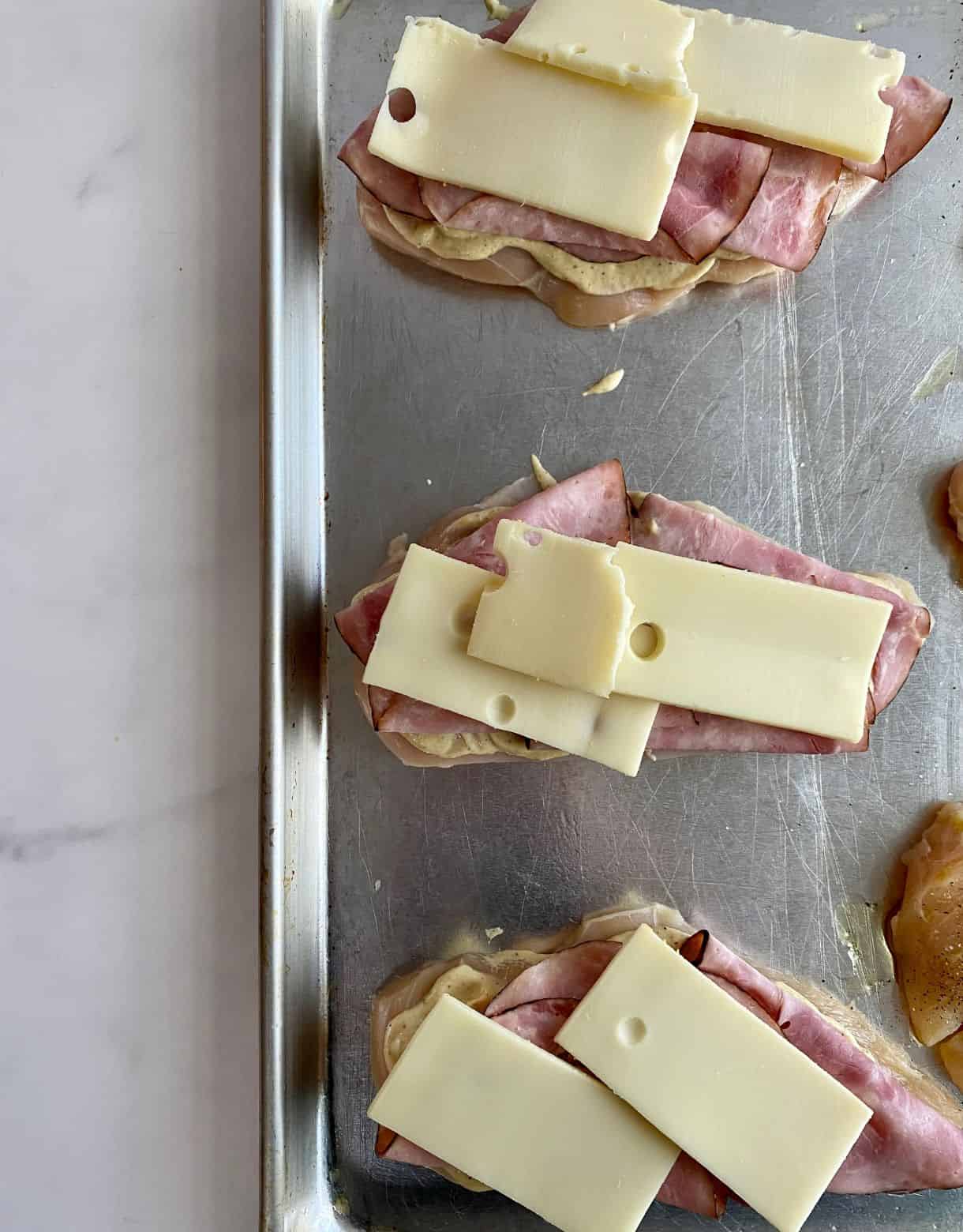 A sheet pan with raw chicken slathered in Dijon sauce, then topped with slices of ham and Swiss cheese.