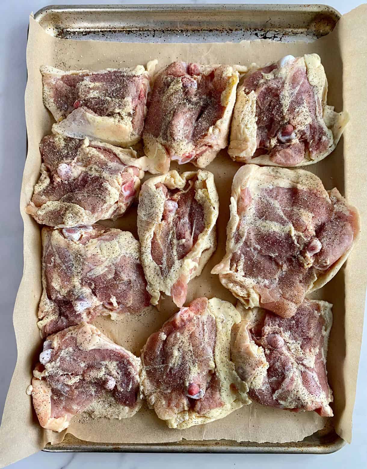 A sheet pan lined with parchment with raw chicken thighs upside down and seasoned with salt and pepper.