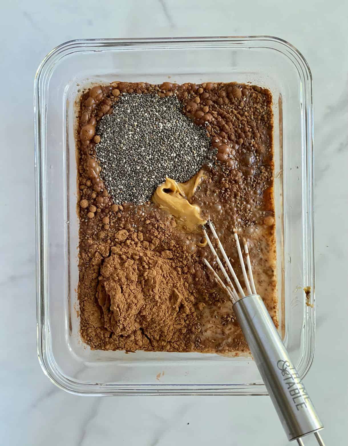 A dish with almond milk, chia seed, cocoa powder, peanut butter, monk fruit sweetener and vanilla all added but not yet mixed.