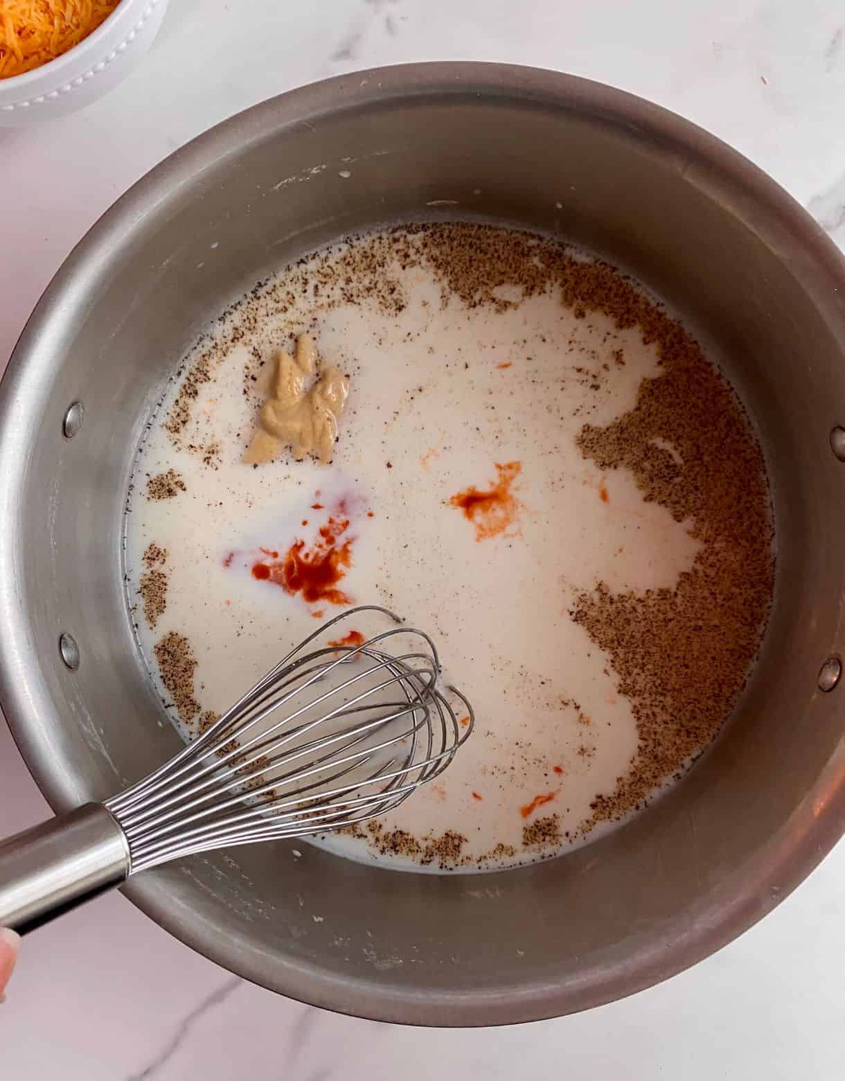 A pot with milk, dijon, hot sauce, salt, pepper and a whisk.
