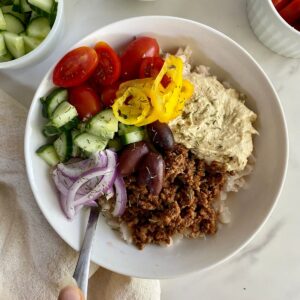 A bowl with cauliflower rice, seasoned grund beef, hummus, kalamata olives, siced red onion, chopped cucumbers, halved grape tomatoes and banana peppers.