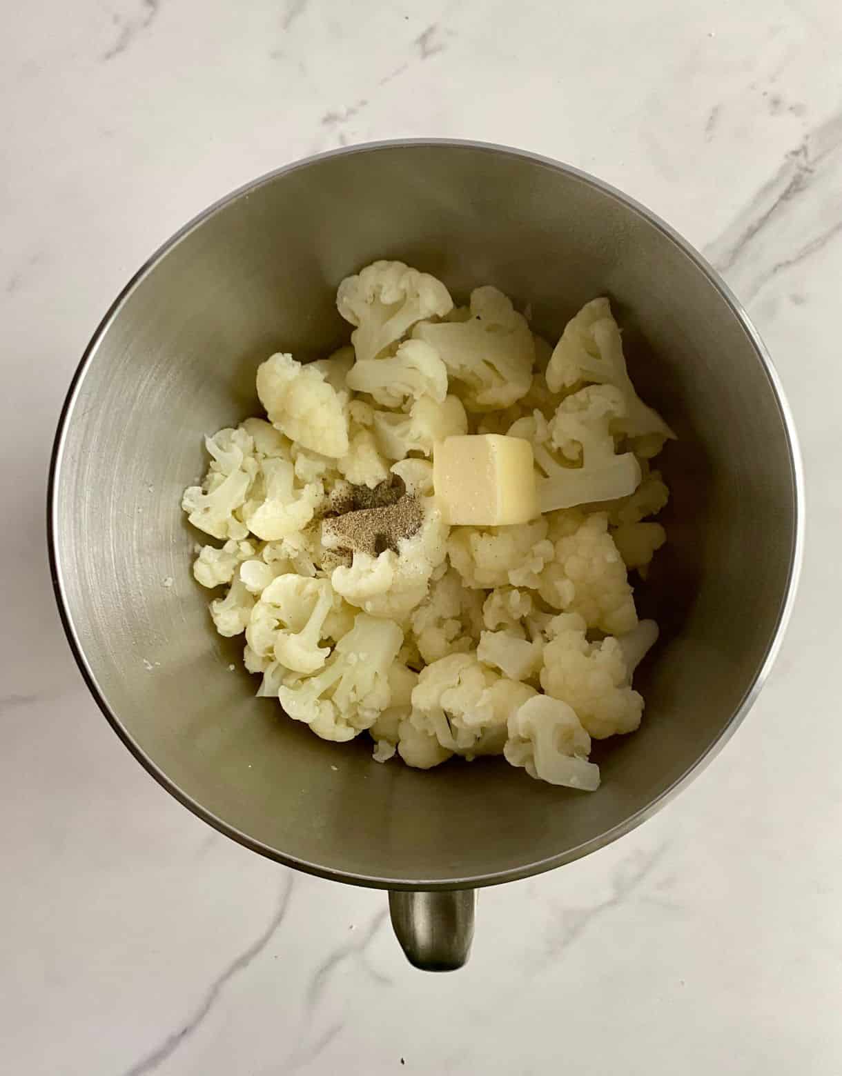 A mixing bowl with cooked cauliflower, butter, salt and pepper.