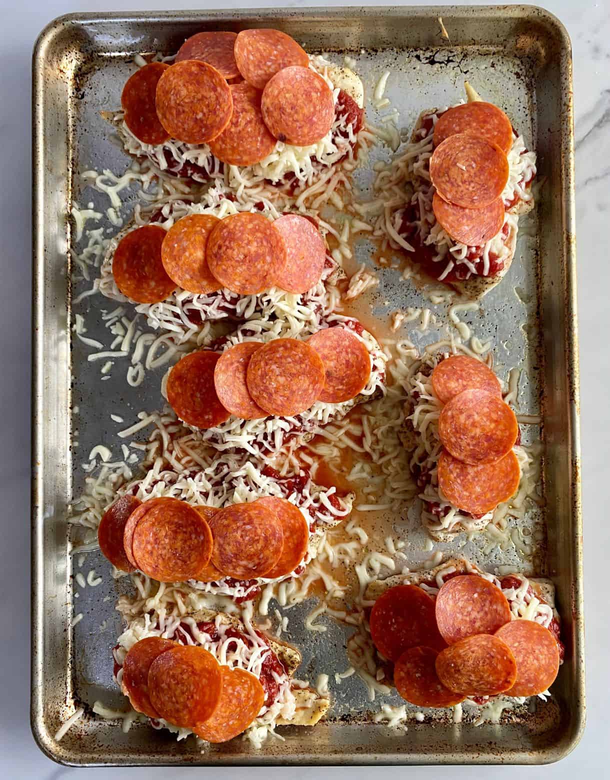 A baking sheet with cooked chicken cutlets topped with marinara sauce, pepperoni and mozzarella and ready to bake.