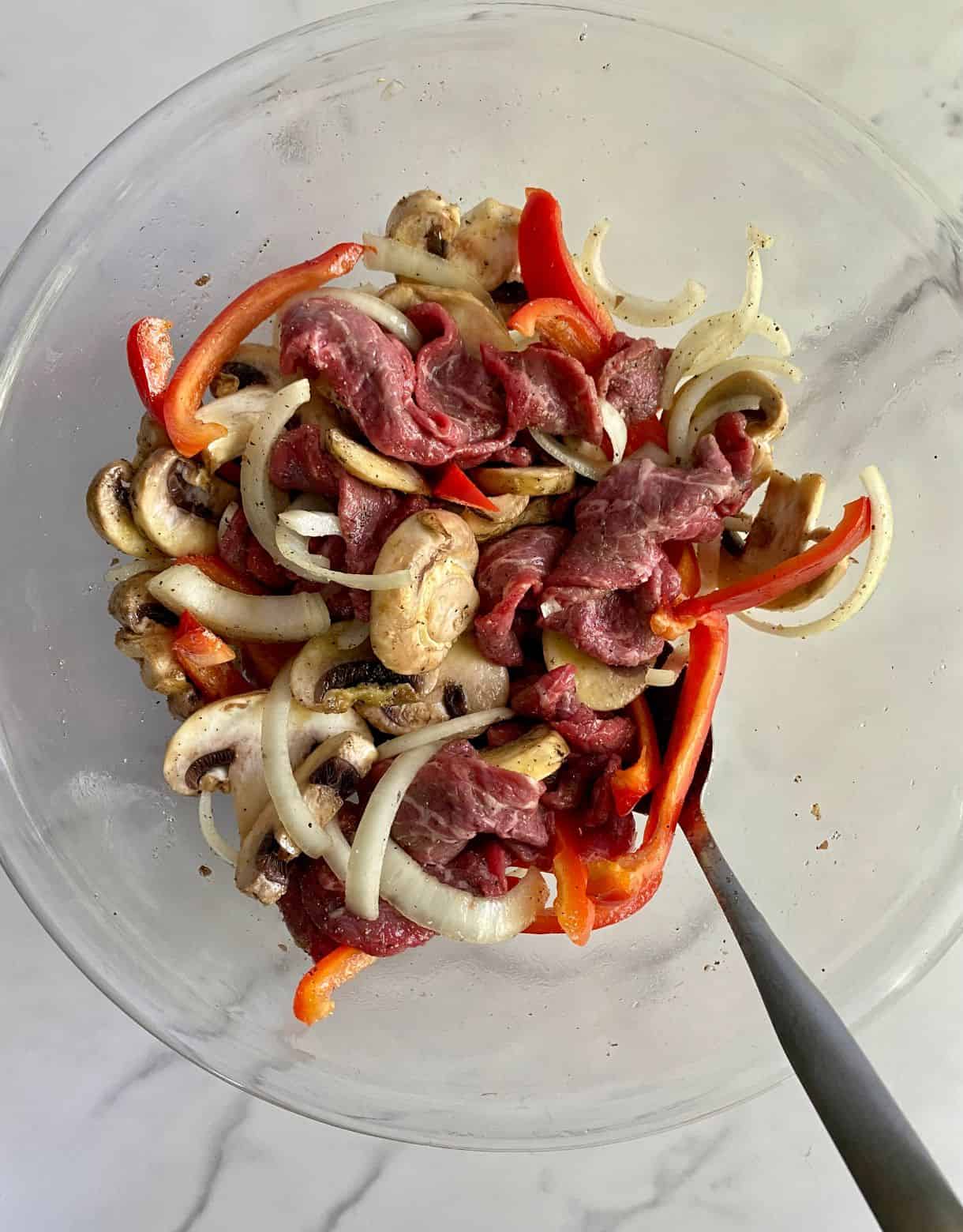 A bowl with sliced steak, onions, bell pepper and mushrooms mixed with olive oil, salt and pepper.