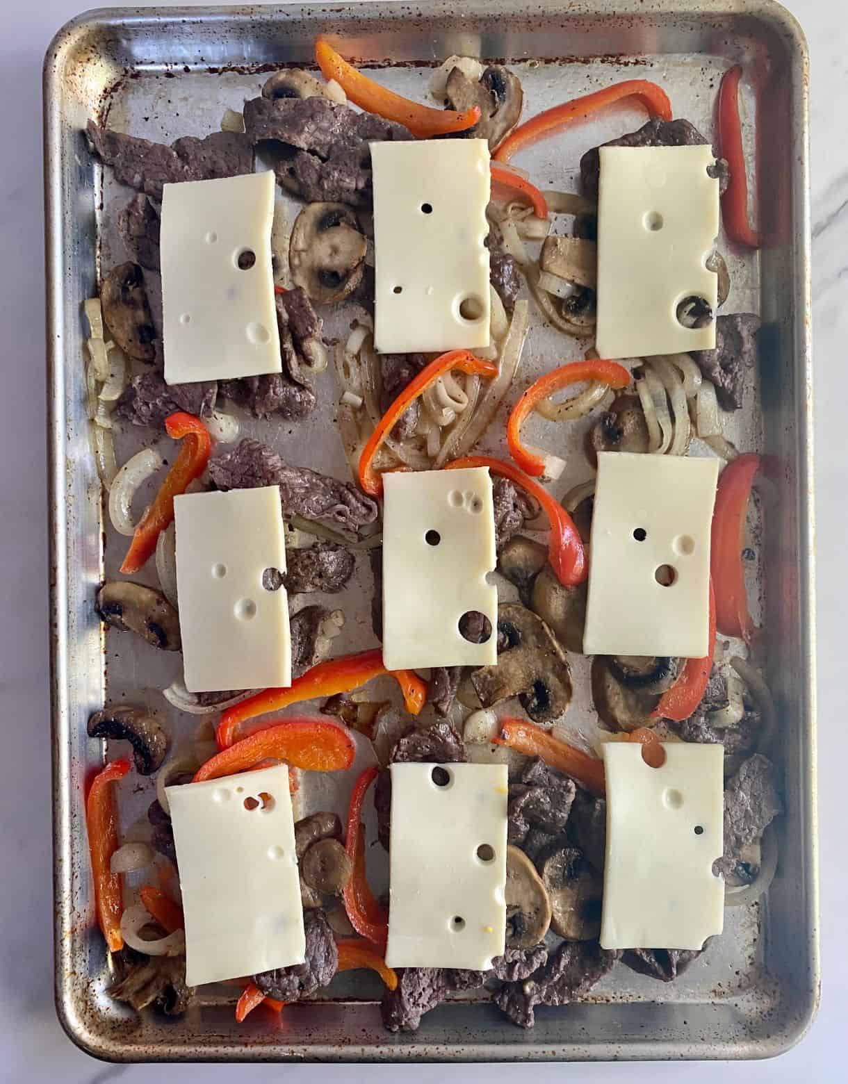 A sheet pan with cooked cheesesteak with onions, mushrooms and peppers topped with Swiss cheese and ready for the final bake to melt the cheese.