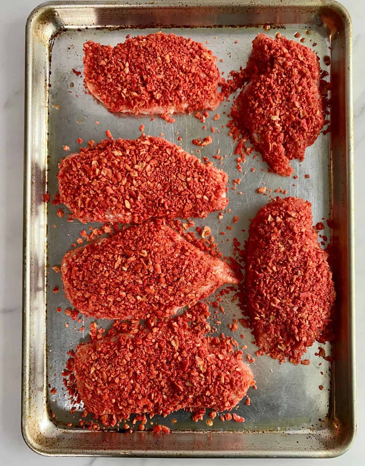 A sheet pan of raw chicken cutlets coated in crushed Takis and ready to bake.