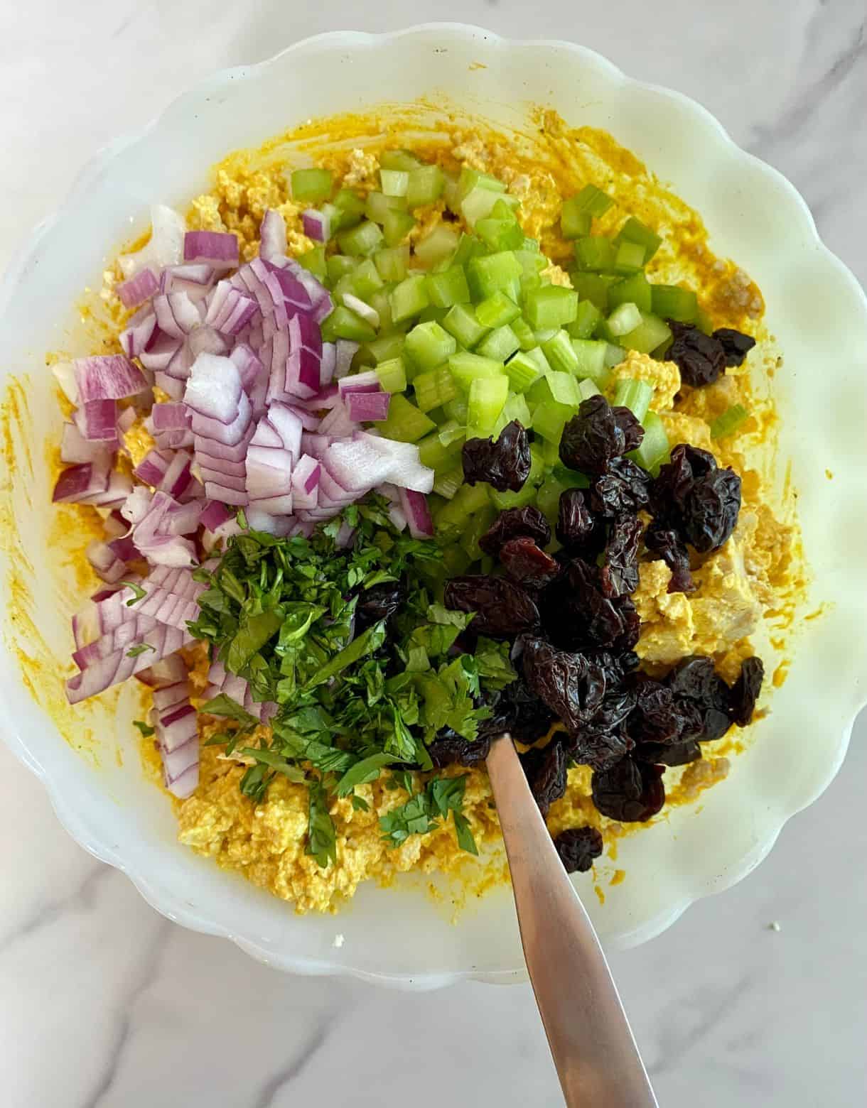 A bowl with curry dressing, crumbled tofu, chopped red onion, chopped celery, diced cilantro and dried cranberries all added and ready to be stirred.