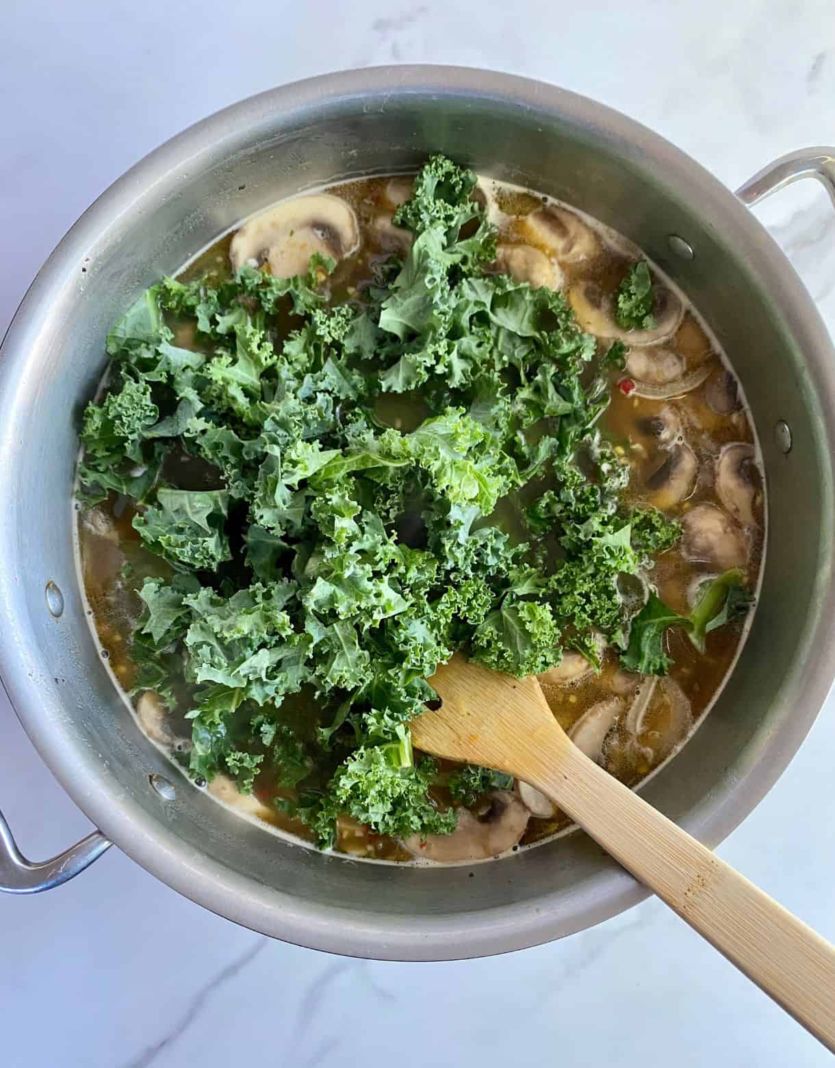 A pot of soup with torn pieces of kale added on top but not yet stirred in.