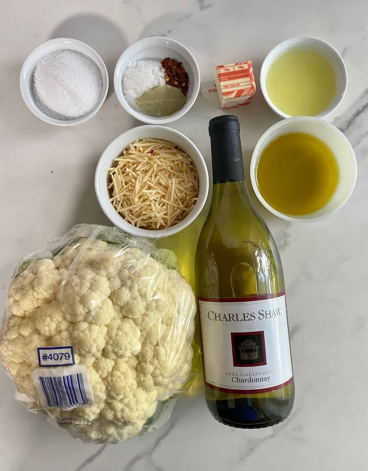 A bottle of white wine, a head of cauliflower, parmesan cheese, olive oil, lemon juice, butter, sugar, salt, red pepper flakes and a bay leaf.